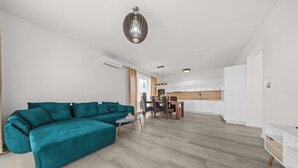 A spacious living area with a teal sectional sofa, a dining table with chairs, and a kitchen in the background.