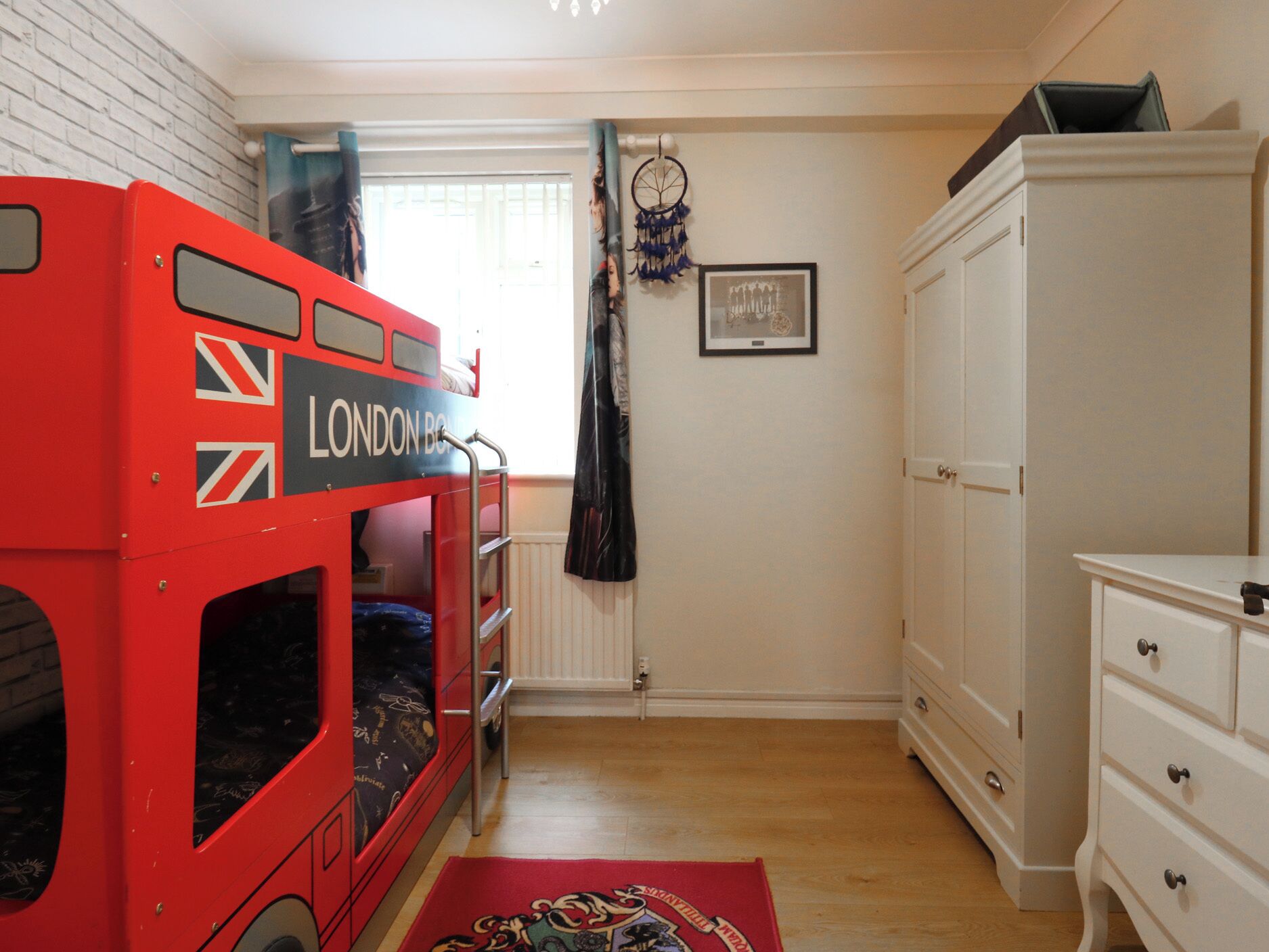 A room with a red double-decker bunk bed labeled "LONDON BUSES," a white dresser, a dreamcatcher, and a framed picture on the wall.