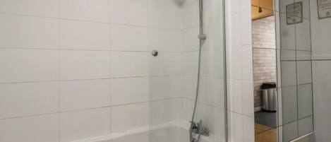 A modern bathroom with a glass-enclosed shower, white tiles, and a visible trash bin.