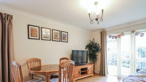 Ein gemütliches Wohnzimmer mit einem runden Holztisch, vier Korbstühlen, einem Flachbildfernseher auf einem Holzschrank und einem großen Fenster mit Vorhängen.