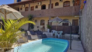A pool area with lounge chairs and umbrellas, a small pool, and a building with multiple windows and balconies. - Hotel Vale do Rio (Sao Goncalo do Abaete)
