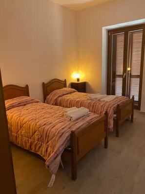 A hotel room with two wooden beds, a nightstand with a lamp, and a door with blinds. - Holiday Home 'Il Focolare dei Sibillini' with Mountain View, Balcony & Wi-Fi (Fematre)