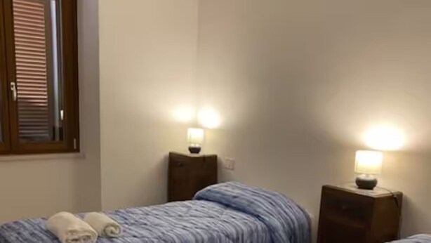 Ein Hotelzimmer mit zwei Betten, jeweils mit blauer Streifenoptik und einem aufgerollten Handtuch. Es gibt zwei Nachttische mit Lampen.