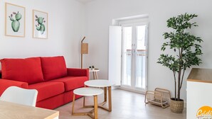 Ein modernes Wohnzimmer mit einem roten Sofa, einem Holzesstisch und einem kleinen weißen Tisch. An den Wänden hängen zwei Bilder von Kakteen in Rahmen, eine Korbstuhl-Stehlampe und ein Topfpflanze stehen dort.