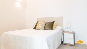 Una cama bien hecha con una colcha blanca y dos almohadones decorativos, una mesita de noche con lámpara, y un enchufe en la pared.