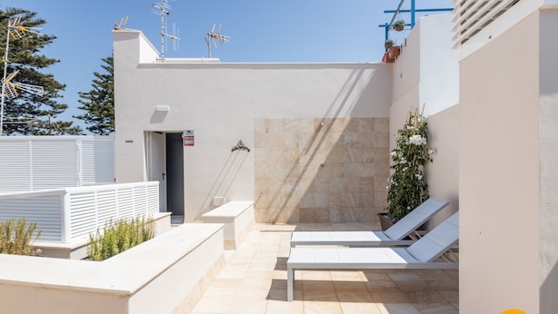 Una terraza en la azotea con dos sillones blancos, una puerta pequeña y una planta en maceta.