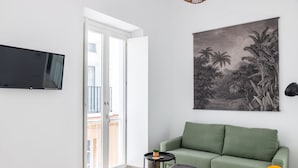 Un salón con un sofá verde, un televisor de pantalla plana, una pequeña mesa con una planta y un cuadro de paisaje grande en la pared.