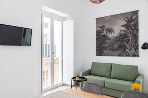 Ein Wohnzimmer mit einem grünen Sofa, einem Flachbildfernseher, einem kleinen Tisch mit einer Pflanze und einem großen Landschaftsbild an der Wand.