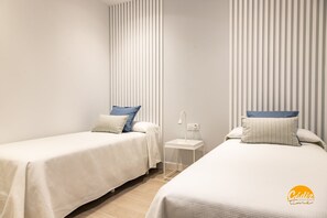 Two single beds with white bedding and blue pillows, a small bedside table with a lamp, and vertical blinds on the windows. - Apartamento Villalobos 1 by C diz Time Apartments (Cádiz)