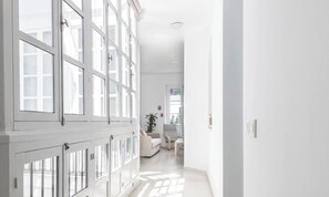 Ein heller, geräumiger Flur mit großen Fenstern, weißer Innenausstattung und Blick in einen weiteren Raum mit Sofa und Pflanze.
