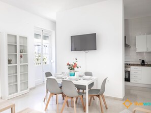 Ein moderner Essbereich mit einem weißen Tisch, gedeckt für vier Personen, grauen Stühlen, einem flachbildfernseher über dem Tisch und einer Küche mit weißen Schränken und einer Arbeitsplatte.