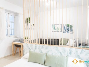 A room with a bed, a desk, and a chair. There is a window with curtains, a plant, and artwork on the wall. - Estudio Palma by Livework (Cádiz)