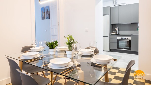 A modern dining area with a glass table, black and white checkered floor, and grey chairs. A kitchen with grey cabinets and a built-in oven is visible in the background. - La Casa del Alfarero by Cadiz Time (Cádiz)