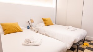 A hotel room with two beds, each with white bedding and a mustard-colored pillow. There is a nightstand with a lamp and a blue cup. A towel is rolled on one bed. - La Casa del Alfarero by Cadiz Time (Cádiz)