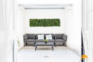 Een moderne buiten zithoek met een grijze bank, twee decoratieve kussens, een klein tafeltje en een groene wandplant.