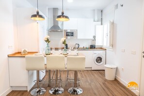 A modern kitchen with white cabinets, a built-in oven, and a washing machine. There are three bar stools and a dining table with a vase of flowers. - El Patio del Mar by Cadiz Time (Cádiz)
