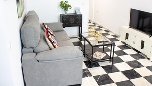 Ein Wohnzimmer mit grauem Sofa, einem schwarz-weißen Schachbrettfußboden, einem Glastisch, einer schwarzen TV-Konsole und einer kleinen Pflanze auf einem Beistelltisch.