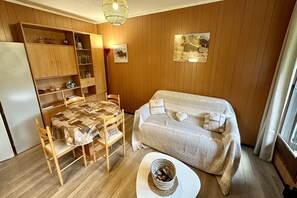 A cozy room with a wooden paneled wall, a dining table with chairs, a sofa, and a small white table with a basket. - Alpine refuge near the forest road (Mont-Dore)