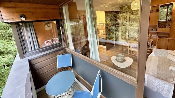 Ein gemütliches Zimmer mit Holzbalkendecke, einem großen Fenster, einem kleinen runden Tisch und einem blauen Stuhl.