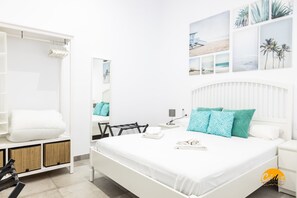 Quarto de hotel moderno com uma cama grande, roupa de cama branca, mesa de cabeceira com abajur e parede decorada com quadros de praia e palmeiras.