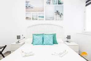 Quarto de hotel com cama branca, duas almofadas a combinar, cabeceira branca, duas mesas de cabeceira com abajures e uma parede com quadros emoldurados.