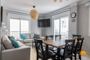 A modern living room with a sofa, a dining table, and chairs. There is a TV mounted on the wall and a clock on the wall. - Casa Arcoiris by Cadiz Time (Cádiz)