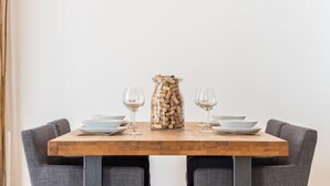 Una mesa de comedor con tapa de madera, rodeada de sillas grises tapizadas. Hay dos copas de vino y una jarra de vidrio transparente llena de tapones de corcho sobre la mesa.