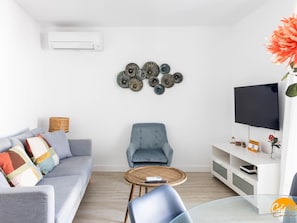 A modern living room with a grey sofa, a blue armchair, a round coffee table, a flat-screen TV, and decorative wall art. - Apartamento Oceano by Cadiz Time (Cádiz)