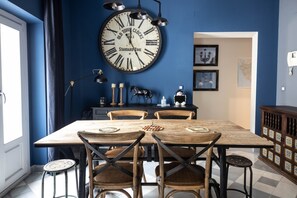 A dining area with a large wall clock, wooden chairs, a table, and a cabinet with decorative items. - Blue Home by Cadiz Time (Cádiz)