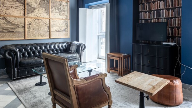 Ein Wohnzimmer mit einem schwarzen Ledersofa, einem Holztisch, einem braunen Ledersessel, einem Glastisch, einem Bücherregal und einer großen Weltkarte an der Wand.