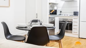 Een moderne keuken met witte kasten, een magnetron, fornuis en een koelkast. Er staat een eettafel met stoelen en een fotokader hangt aan de muur.