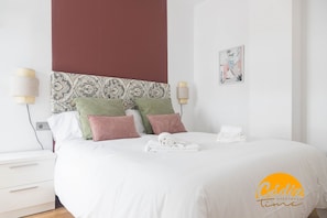 A neatly made bed with white linens and a rolled towel, a bedside table with a lamp, and a wall-mounted light fixture. - Apartamento Caleta Free Parking by Cadiz Time (Cádiz)