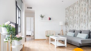 Ein modernes Wohnzimmer mit grauem Sofa, weißem Couchtisch und einem Korbstuhl. Eine weiße Regalanlage beherbergt Bücher und eine Pflanze, und an der Wand hängt ein großes abstraktes Kunstwerk.