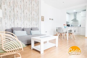 A modern living room with a grey sofa, a white coffee table, and a wicker chair. A kitchen with white cabinets and a grey countertop is visible in the background. - Apartamento Caleta Free Parking by Cadiz Time (Cádiz)
