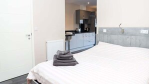 Ein Hotelzimmer mit einem Bett, einem ordentlich bezogenen Bett mit Handtuch, einer weißen Tür und Blick in den Küchenbereich mit Schränken und Geräten.