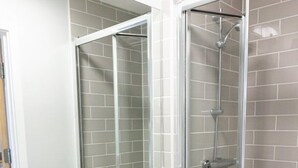 A bathroom with a white sink, a mirror, a shower with a glass door, and a trash bin. - Chic Studio in Cheltenham (Cheltenham)