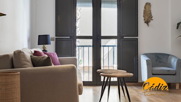 A living room with a sofa, a small round table, a chair, and a large window with blinds. - Corazon del Mar Free Parking by Cadiz Time (Cádiz)