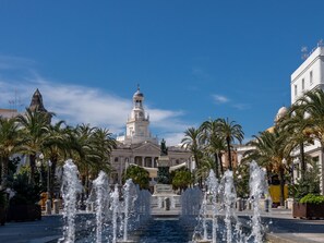 A fountain with water jets in a plaza, surrounded by palm trees and a large building with a clock tower. - La Luz de Cadiz by Cadiz Time (Cádiz)