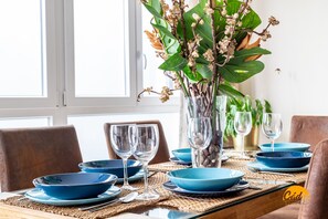 Ein Esstisch ist gedeckt mit blauen Tellern, klaren Gläsern und einer Vase mit grünen Blättern und Blumen.