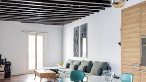 A modern living room with a glass dining table, teal chairs, a wooden coffee table, and a sofa with grey cushions. There is a wooden cabinet with a built-in oven, and a large window with a view of the city. - Diana de Columela by Cadiz Time (Cádiz)