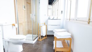 A modern bathroom with a glass shower enclosure, a white toilet, a wooden bench with towels, and a white sink with a mirror above it. - Diana de Columela by Cadiz Time (Cádiz)