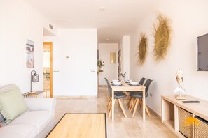 A modern living room with a sofa, dining table, and chairs. There is a TV mounted on the wall and a small table with a lamp. - Casa Capuchinos Free Parking by Cadiz Time (Cádiz)