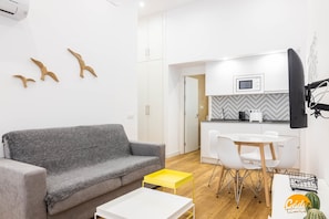 A modern living room with a grey sofa, a yellow side table, and a white coffee table. A kitchen with white cabinets and a microwave is visible in the background. - Estudio Gaviota by Cadiz Time (Cádiz)