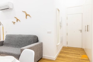 A room with a grey sofa, a white chair, a small round table, and a white door with a phone mounted on it. - Estudio Gaviota by Cadiz Time (Cádiz)