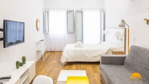 A modern hotel room with a bed, a sofa, a TV, a desk with a computer, and a small plant. - Estudio Gaviota by Cadiz Time (Cádiz)