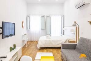 A modern hotel room with a bed, a sofa, a TV, a desk with a computer, and a small plant. - Estudio Gaviota by Cadiz Time (Cádiz)