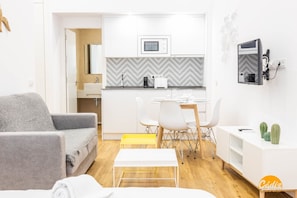 A modern living room with a grey sofa, a small white table, and a yellow bench. There is a kitchen area with white cabinets, a microwave, and a coffee machine. A TV is mounted on the wall, and there are two green cacti on a white shelf. - Estudio Gaviota by Cadiz Time (Cádiz)
