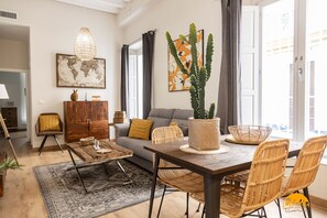 Uma sala de estar moderna com sofá, mesa de jantar, cadeiras de vime, mesa de centro, uma obra de arte com mapa mundi, uma planta de cacto e uma janela grande com cortinas.