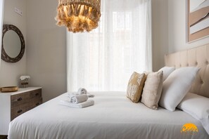 Uma cama bem arrumada com lençóis brancos, uma mesa de cabeceira de madeira com uma tigela e flores, e um abajur pendente.