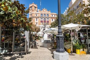 Uma rua de paralelepípedos com cafés ao ar livre, uma fonte central e um edifício histórico ao fundo.